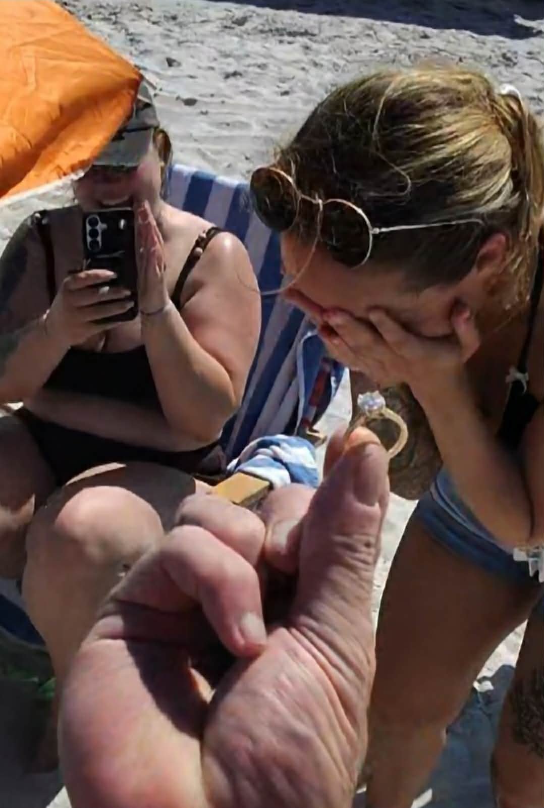 Two women on a beach, one is holding a smartphone and appears to be surprised or excited, while the other is covering her face with her hand and leaning forward. A large hand holding a lighter is in the foreground, close to the woman with sunglasses.