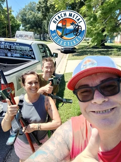 Three people taking a selfie outdoors next to a vehicle with a sign for Beach Metal Detectives. The person in the foreground wears sunglasses and a red cap, while the other two hold metal detectors, smiling at the camera.