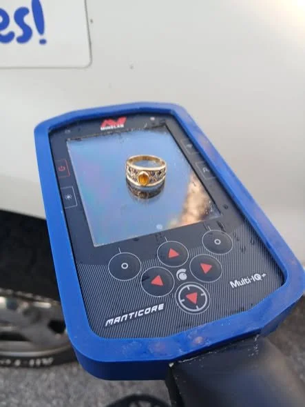 A gold ring with a gemstone on top sits on a device that looks like a metal detector or a diagnostic scanner.