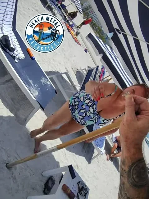 Woman on a beach with striped umbrellas, holding a beach spoon towards the camera, with a tattooed arm reaching out. Items like sunglasses, a speaker, and a brochure are on the sand. A logo for Beach Metal Detectives is visible.