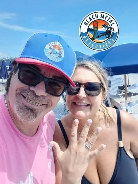 A smiling man and woman at the beach, both wearing sunglasses and casual summer clothes, with beach umbrellas and chairs in the background. The man is wearing a pink shirt and a blue cap with a beach-themed logo, and the woman is showing off her ring.