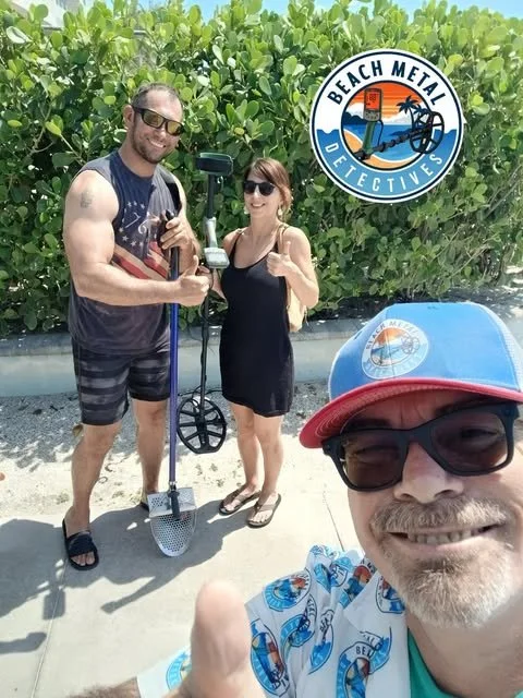 Three people taking a selfie at the beach, with a man in the foreground wearing sunglasses and a hat, and a couple behind him holding a metal detector. A sign reading 'Beach Metal Detectives' is visible in the background.