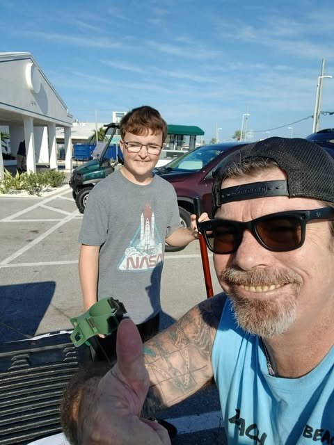 A man wearing sunglasses and a backward cap taking a selfie with a boy in a parking lot, both smiling. The man is giving a thumbs up, and the boy is wearing a NASA t-shirt. There are parked cars and a building in the background under a partly cloudy sky.