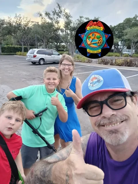Group of four people, two children and two adults, taking a selfie in a parking lot with trees in the background. The man in the foreground is wearing glasses and a hat, giving a thumbs-up. The woman and children are smiling, and one child is holding a toy gun.