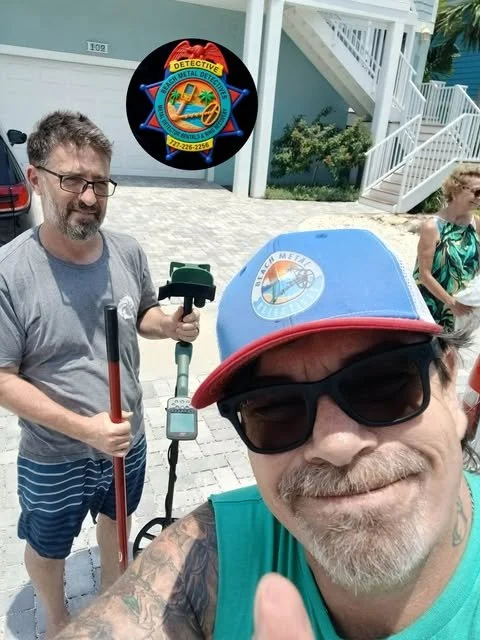 A man taking a selfie with two people in the background, one of whom is holding a metal detector. The man in the foreground is wearing sunglasses and a colorful cap, smiling. The background shows a driveway, stairs, and a woman wearing a green dress.