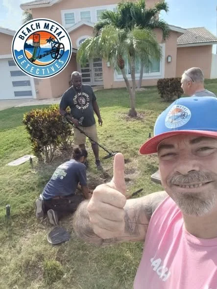 A man in a pink shirt wearing a Beach Metal Detective hat takes a selfie with a thumbs-up, while two other men and a woman work together in the garden of a house with a palm tree. One man is standing with a metal detector, and the others are kneeling or sitting near the ground, possibly digging or examining something.