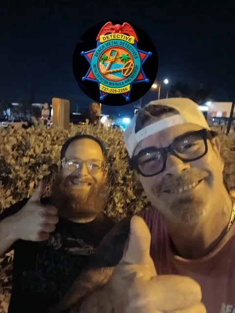 Two men are outdoors at night, smiling and giving thumbs-up gestures. One man is wearing glasses and a cap, the other has glasses and a full beard. In the background, there is a sign with a sheriff badge shape that reads 'Beach Metal Detectives' along with a phone number and other contact info. The scene appears to be at a casual outdoor event or gathering.