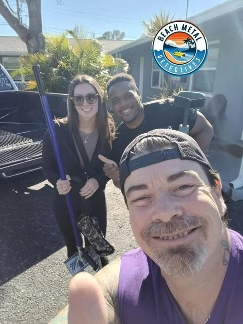 Three smiling people taking a selfie outdoors in a driveway on a sunny day, with a pickup truck behind them. The group includes a woman with sunglasses, a man with a cap, and another man pointing at the camera. There is a logo for 'Beach Metal Detectives' in the top right corner.