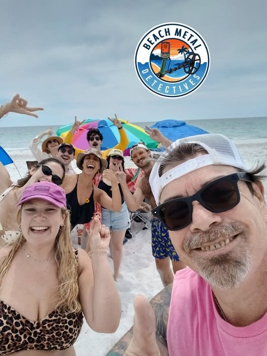 A group of people on a beach, smiling and posing for a selfie, with ocean and cloudy sky in the background, featuring a Barbados beach rescue crew member, beach umbrellas, and the logo of Beach Media Detectives.