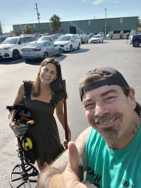 A man and a woman taking a selfie in a parking lot with cars and a large building in the background. The woman is smiling and holding a bicycle, while the man, wearing a cap backwards, is giving a thumbs-up.