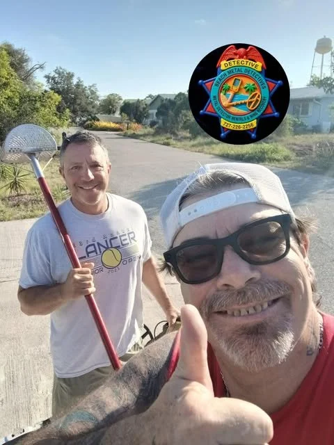 Two men taking a selfie outdoors on a sunny day. The man in the foreground is wearing glasses, a cap backward, and a red shirt, giving a thumbs-up. The man in the background is holding a tennis racket and smiling. A badge with a badge logo for a metal detector is floating in the air above them.