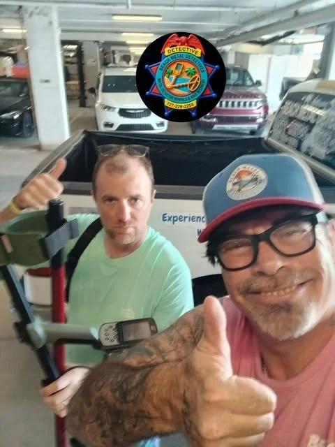 Two men taking a selfie in a parking garage. The man in the foreground wears glasses, a baseball cap, and a pink T-shirt, giving a thumbs-up. The man in the background wears a light green shirt, with a walker nearby, and is pointing at the camera with a serious expression. Several parked cars are visible in the background.