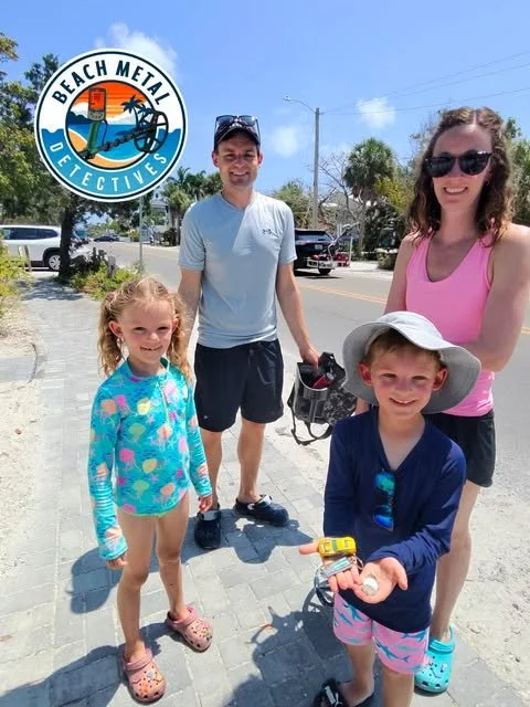 A family of four standing on a sidewalk outdoors with a beach town background. There is a man, woman, and two children, a girl and a boy, smiling. The boy is holding a shell and a small yellow toy. The woman is wearing sunglasses and a pink tank top, and the girl is in a colorful bathing suit. The logo of Beach Metal Detectives is in the upper left corner.