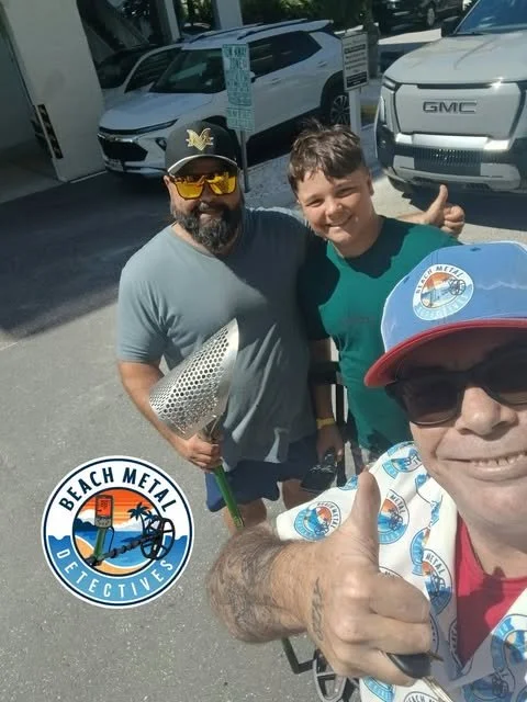 Three men outdoors taking a selfie, one wearing a red cap and sunglasses, the other with a beard and sunglasses, and the third in a teal shirt. Two parked cars and parking signs are in the background. The man with the cap is giving a thumbs up. There is a logo for Beach Metal Detectives on the photo.