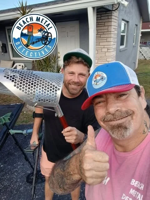 Two men wearing hats, one with a pink shirt giving a thumbs-up, in front of a building, with a metal detector and a scoop, and a logo with palm trees and the text "Beach Metal Detectives."