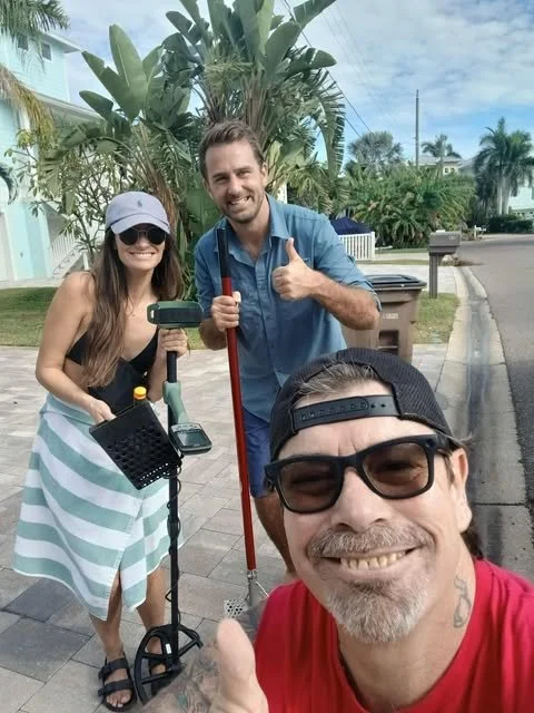Three people smiling and taking a selfie outdoors on a sidewalk with palm trees in the background, one person holding a metal detector.
