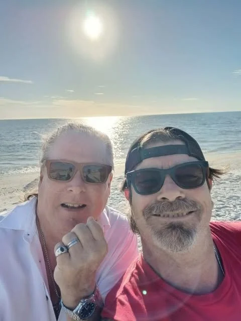 A man and woman taking a selfie on the beach during sunset, both wearing sunglasses, with the ocean and sun in the background.