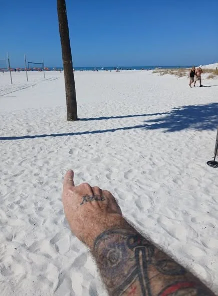 A person with tattoos on their arm pointing at a wooden pole on a sunny beach with white sand and a clear blue sky. Some people are walking in the background, and the ocean is visible in the distance.