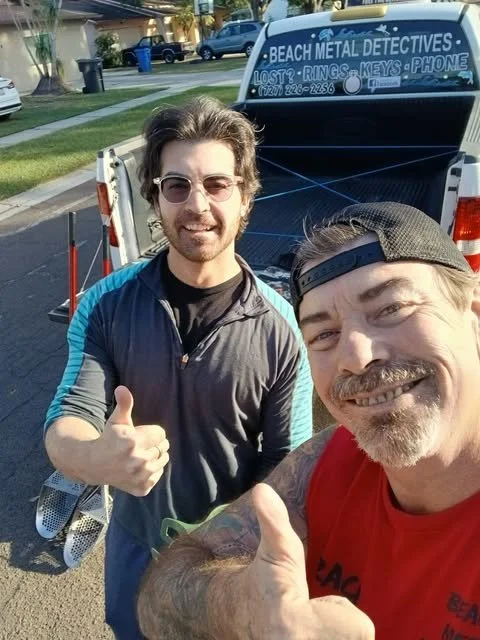 Two men taking a selfie in front of a pickup truck with a sign that reads 'Beach Metal Detectives' and lists services like lost rings, keys, and phones, with a suburban neighborhood in the background.