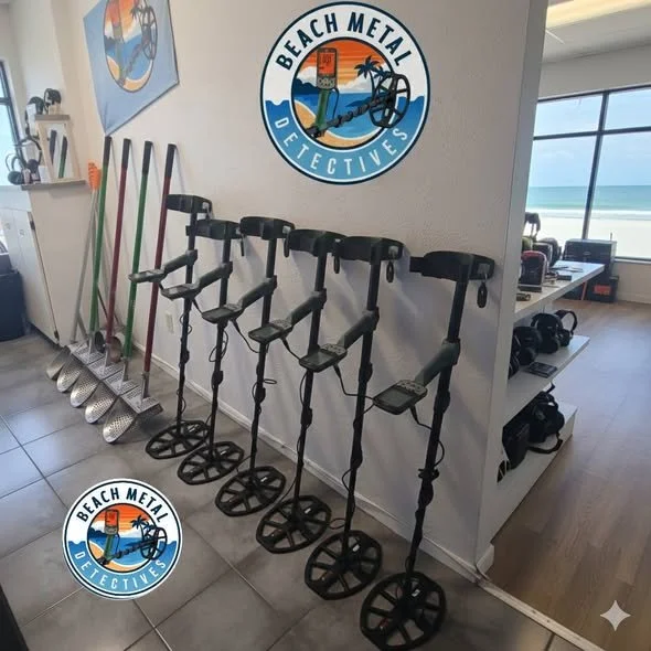 Indoor scene with six metal detectors lined up against a wall with a 'Beach Metal Detectives' logo. There are six beach or metal detecting shovels racked vertically behind the detectors. A large window shows a view of the beach and ocean. Shelves with accessories and a desk are visible on the right.