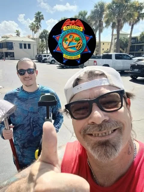 Two men taking a selfie outdoors, one smiling in the foreground wearing sunglasses and a baseball cap, holding a sledgehammer; the other smiling in the background holding a broom. Palm trees and parked cars are visible in the background. A logo for Beach City Metal Detectors is superimposed at the top center of the image.