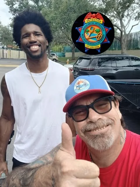 Two men smiling outdoors, one giving a thumbs up. The man in the foreground wears glasses, a red t-shirt, and a blue cap with a logo. The man in the background has curly hair, a white sleeveless shirt, and a gold chain. There is a badge/logo at the top of the image.