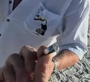 A person wearing a white shirt with a ladybug design on it, holding a pen or marker, possibly signing or writing on something near a gravel surface.