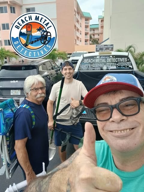 Three men standing in front of a vehicle with a sign that reads 'Beach Metal Detectives' in a parking lot. The man in the foreground is smiling and giving a thumbs up, wearing a red cap and glasses. The middle man has white hair and glasses, and the man in the background is holding a metal detector and smiling.