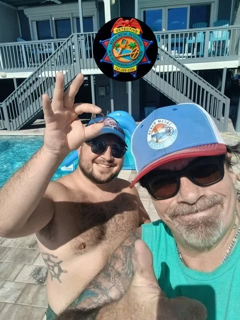 Two men in hats and sunglasses taking a selfie by the pool at a hotel or apartment complex. One man is shirtless, smiling, and making an OK sign with his fingers. The other man is wearing a teal t-shirt, a baseball cap with a logo, and smiling. In the background, outside, are motel-style buildings and a water slide. Above them, a sign for a metal detective agency is visible.