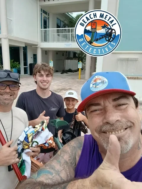 A group of four people smiling for a selfie outside a building with a sign that reads "Beach Metal Detectives" and a logo featuring a metal detector, waves, and a palm tree.