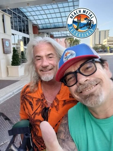 Two smiling men taking a selfie in front of the Beach Metal Detectives logo outside a building.