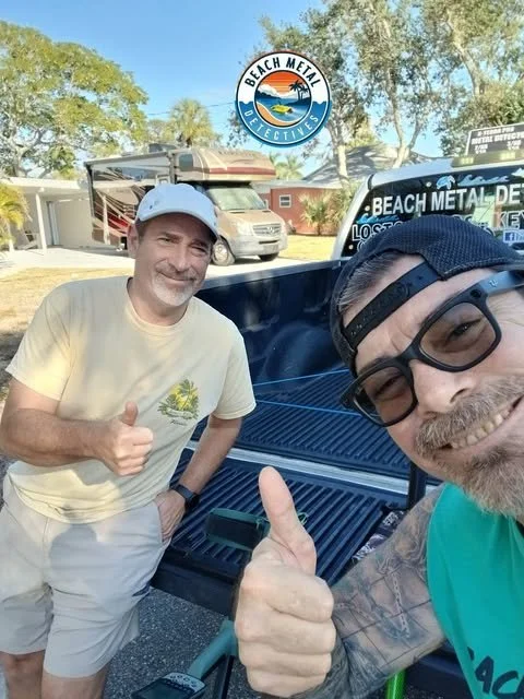 Two men taking a selfie outdoors next to a pickup truck with a Beach Metal Detectives logo. One man is giving a thumbs-up and the other is smiling at the camera.