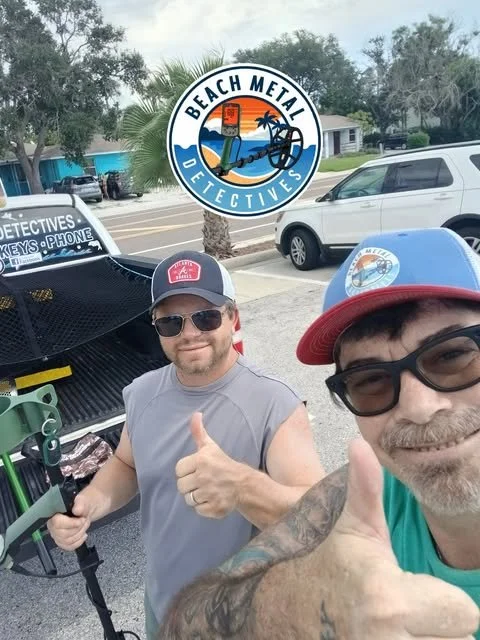 Two men taking a selfie outdoors in a parking lot. One is wearing sunglasses and a cap, giving a thumbs-up. The Other is wearing a hat and glasses, also giving a thumbs-up. A black vehicle with detective signage and equipment is behind them. The logo for Beach Metal Detectives is at the top center, showing a beach scene with a metal detector, palm trees, and a sunset.