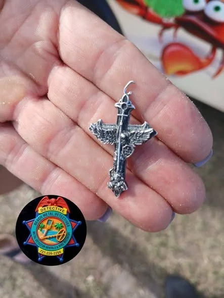 A hand holding a silver-colored necklace with a cross and wings pendant. There is a colorful cartoon-like lobster toy and a badge with a badge with a detective emblem in the background.