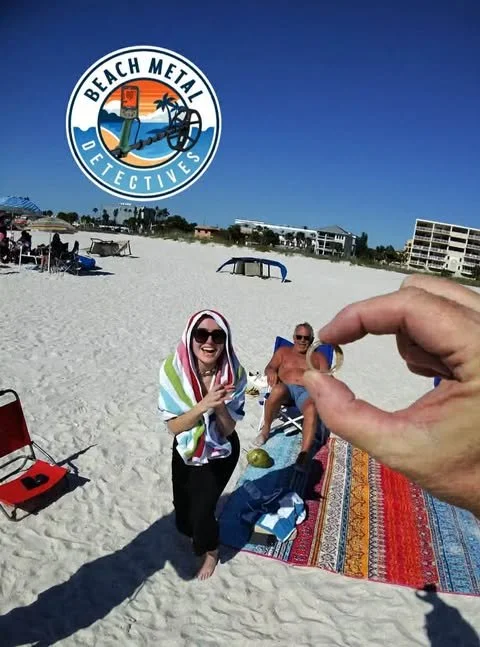 A beach scene with two people sitting on the sand, one woman standing with a towel on her head, and a hand holding a ring close to her. There is a beach towel, a beach chair, and a red speaker. Buildings are visible in the background. The image features the Beach Metal Detectives logo in the top left corner.