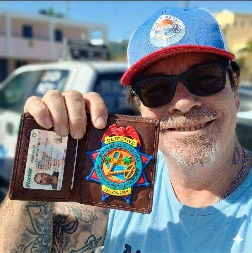 A man wearing sunglasses and a blue cap with a "Black Metal" patch smiles and shows off a brown wallet containing a Florida ID card and a badge for Beach Metal Detectives, which has a colorful design with an anchor, palm trees, and a ring around it. The man has tattoos on his arm and has a background with a white vehicle and buildings.