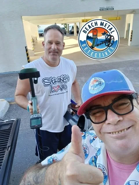 Two men taking a selfie in front of a parking garage with a 'Beach Metal Detectives' logo. One man is smiling and giving a thumbs up, wearing a cap and glasses. The other man is holding metal detecting equipment and smiling.