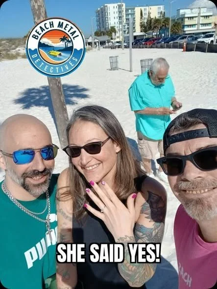 Three people taking a selfie on the beach with a man in the background looking at his phone. A sign with the logo for Beach Metal Detectives is visible. The caption reads 'She Said Yes!'