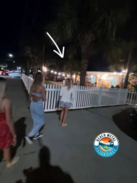 People walking on a sidewalk at night near a white picket fence, with trees and a lit building in the background, and a logo for Beach Metal Detectives.