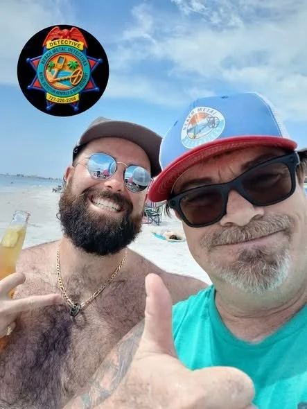 Two men at the beach taking a selfie, smiling, one holding a drink, wearing sunglasses and a hat, with a beach and cloudy sky in the background.
