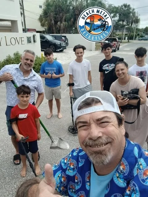 Group of six people (including a man holding a shovel) smiling outdoors in front of a building with palm trees, with a Beach Metal Detectives logo in the sky above.