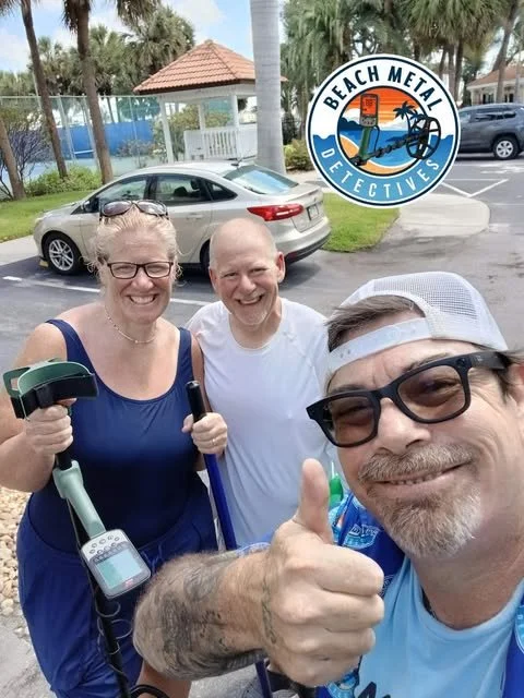 Three smiling people taking a selfie outdoors, with a woman holding a metal detector and two men, one giving a thumbs-up. There is a parked silver car, palm trees, and a logo for Beach Metal Detectives in the background.