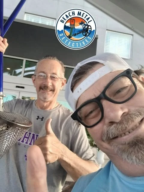 Two smiling men, one giving a thumbs-up, are taking a selfie in front of a