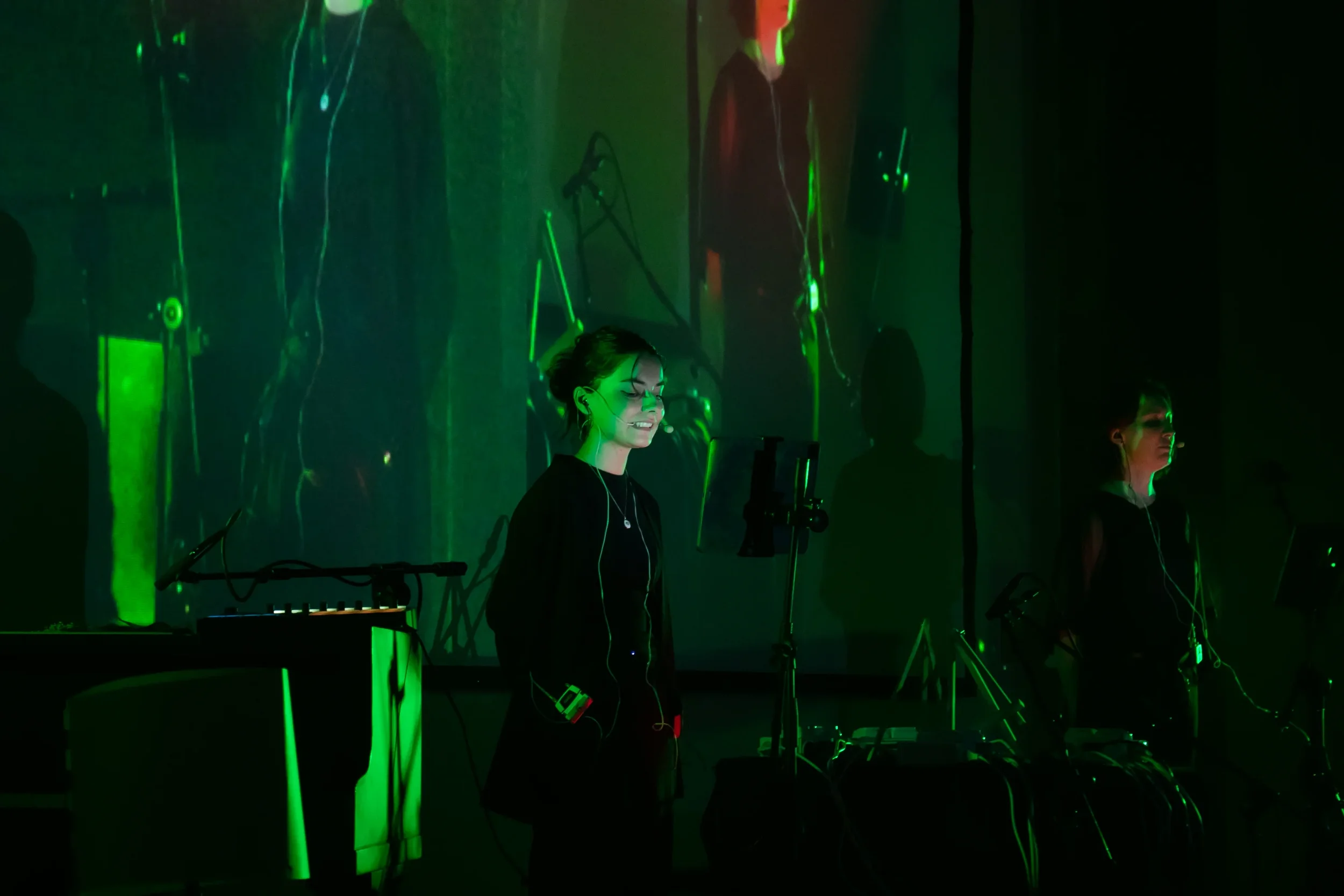 Two women stand in front of a dark background illuminated by green lights, with electronic music equipment around them, one smiling with eyes closed and wearing earphones.