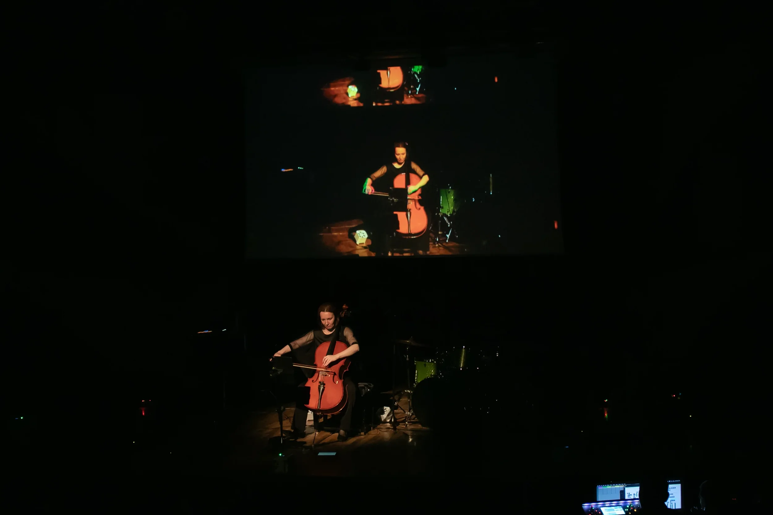 A woman playing a cello on stage with a drum set behind her, visible in a reflection projected on a screen behind her.