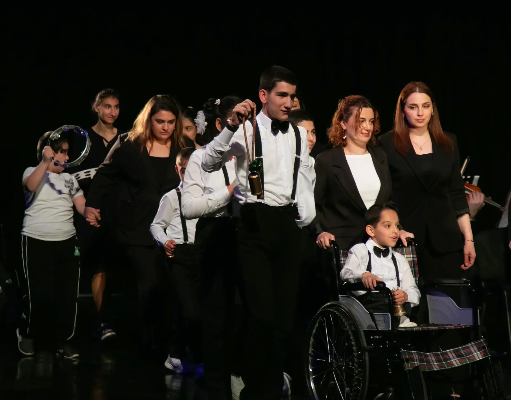 Group of people, including children and adults, standing on stage during a performance or award ceremony. One boy in a wheelchair is in the front, and a tall young man dressed in a white shirt with suspenders holds a medal, with others wearing formal