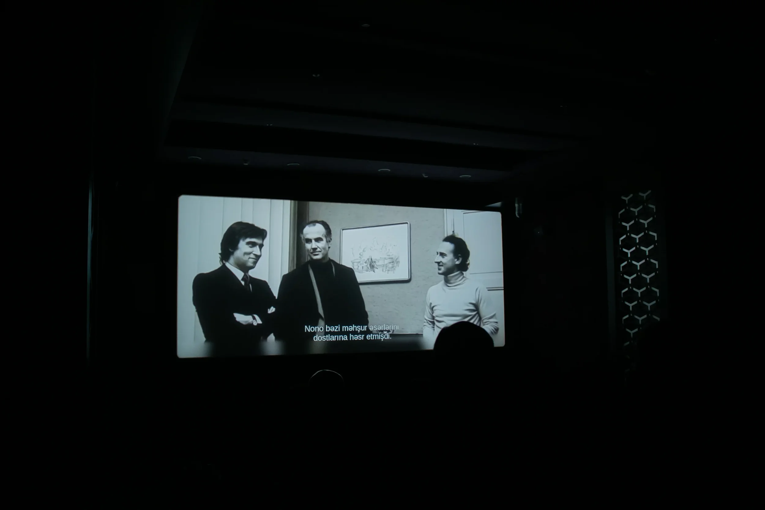 A black and white photo of three men in conversation, with one man on the left crossing his arms, in an indoor setting with a framed picture on the wall behind them. Subtitles in a non-English language are visible at the bottom.