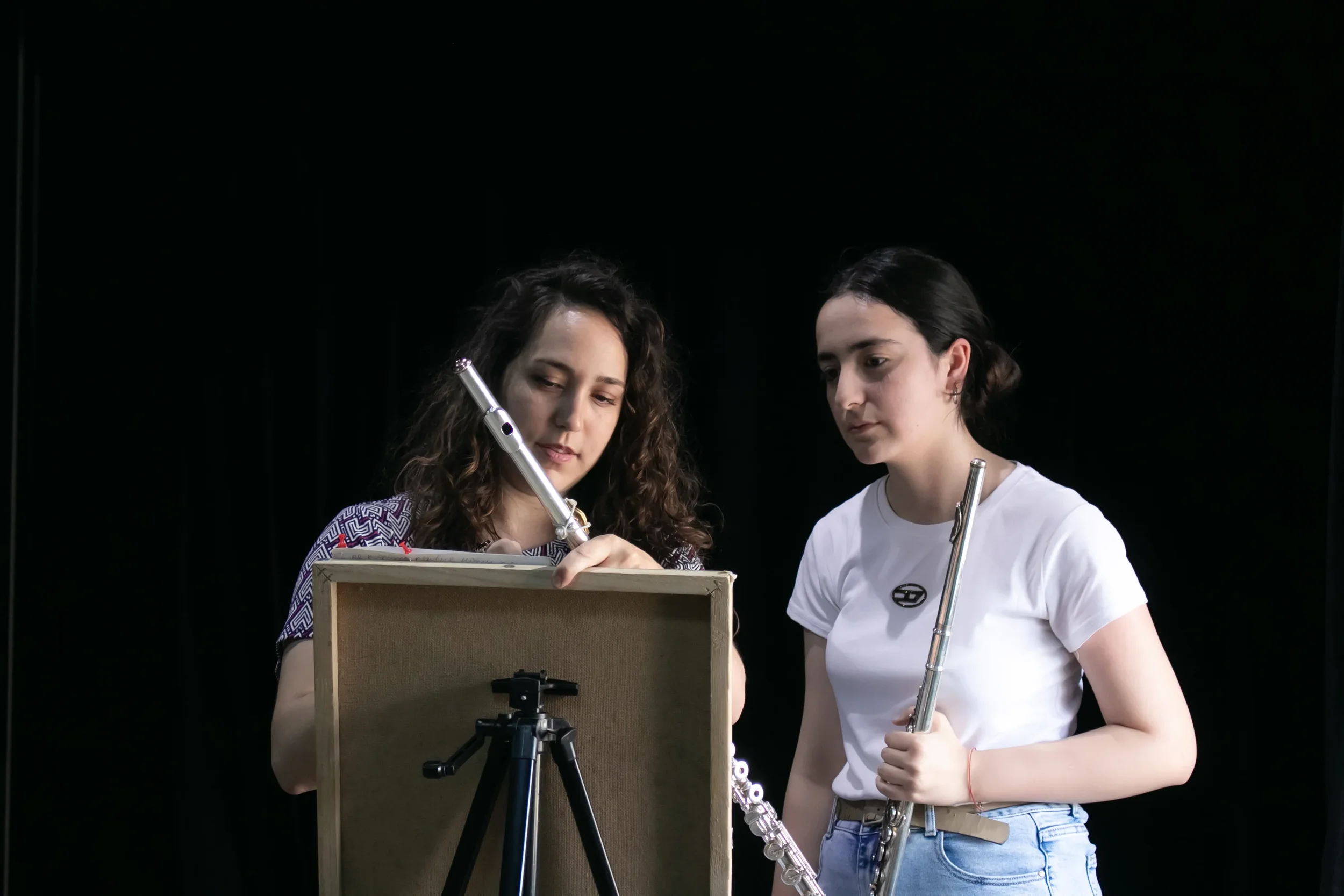 Two women practicing flute playing in a studio with a black background, one adjusting her flute while the other looks on.