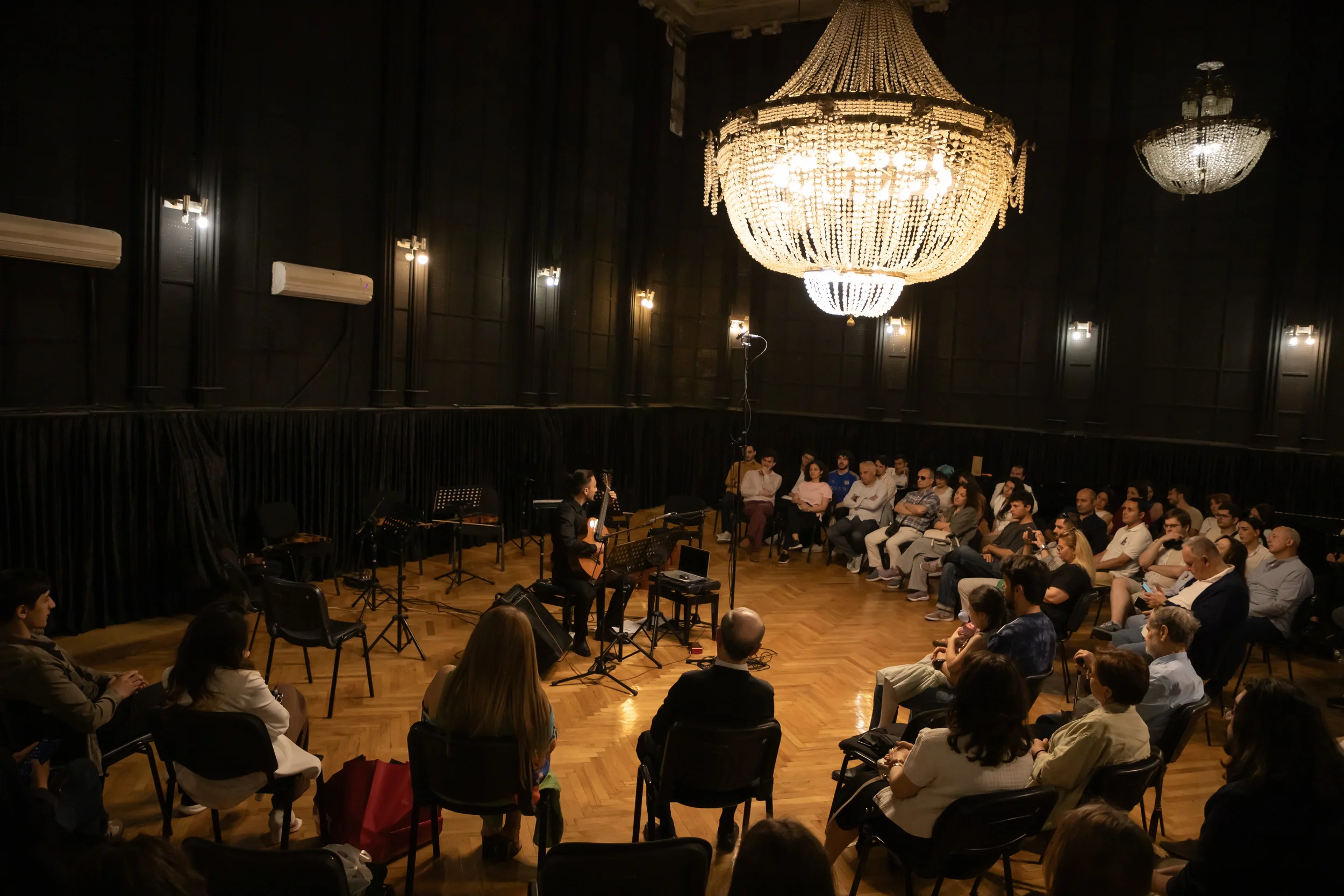A musical performance in an elegant, dimly lit room with a large chandelier. A musician is playing a guitar on stage while an audience listens attentively.