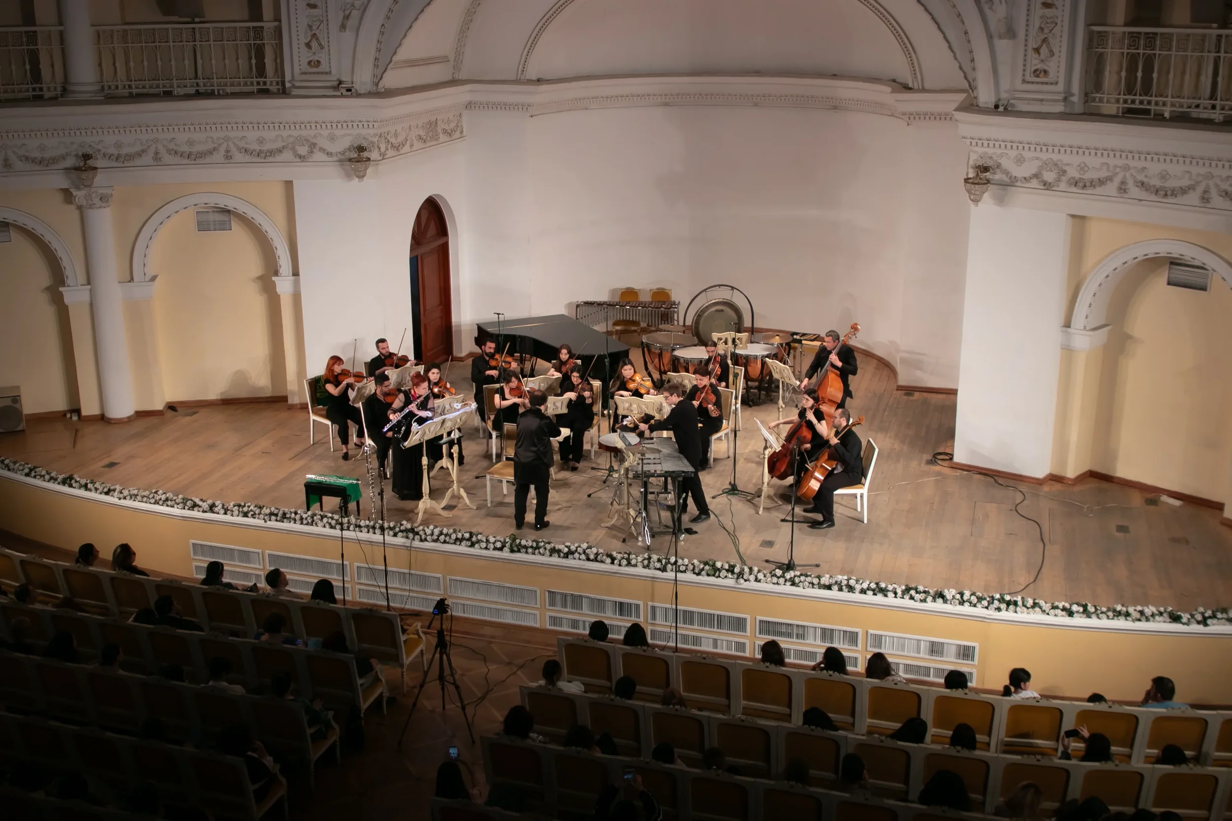 An orchestra performing on stage in a concert hall, with conductor, string, percussion, and other classical instruments, audience seated in front.